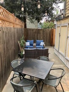 A long patio area. Along a fence there is a square dining table and 4 chairs with a wicker seating area behind it. There are blue pillows on the patio furniture. 